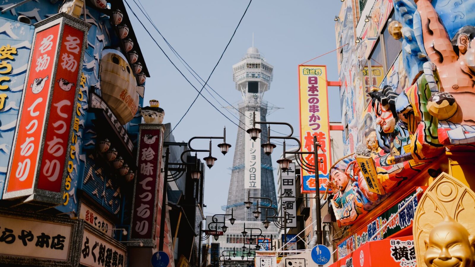 Lively street view in Osaka with vibrant shops and the iconic Tsutenkaku Tower in the background.