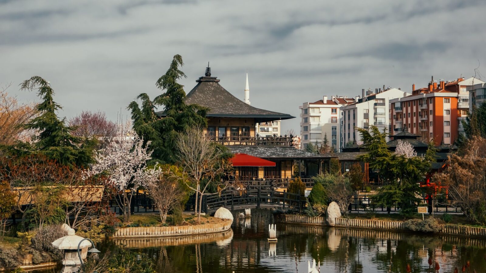 A tranquil Japanese-inspired park in Konya featuring a traditional building, pond, and urban backdrop, perfect for travel enthusiasts.