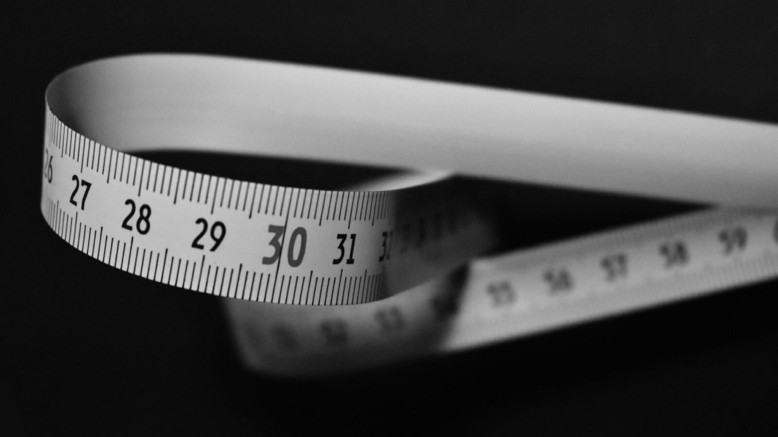 Black and white image of a flexible measuring tape curling on a dark background.